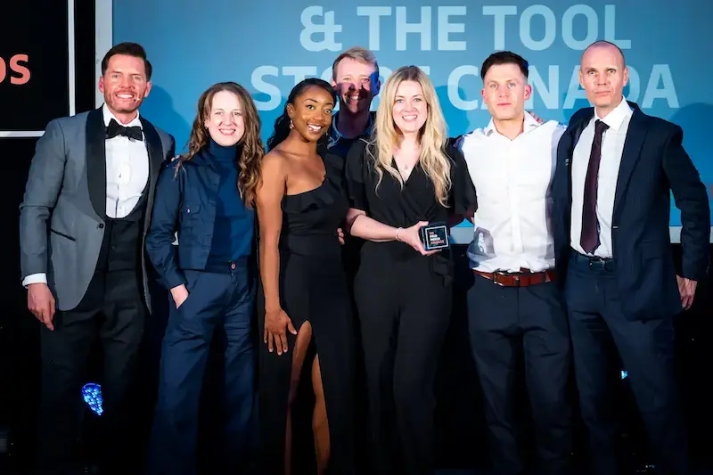 Seven people in formal evening wear posing on stage with an award trophy in front of a blue backdrop reading "& The Tool Store Canada"